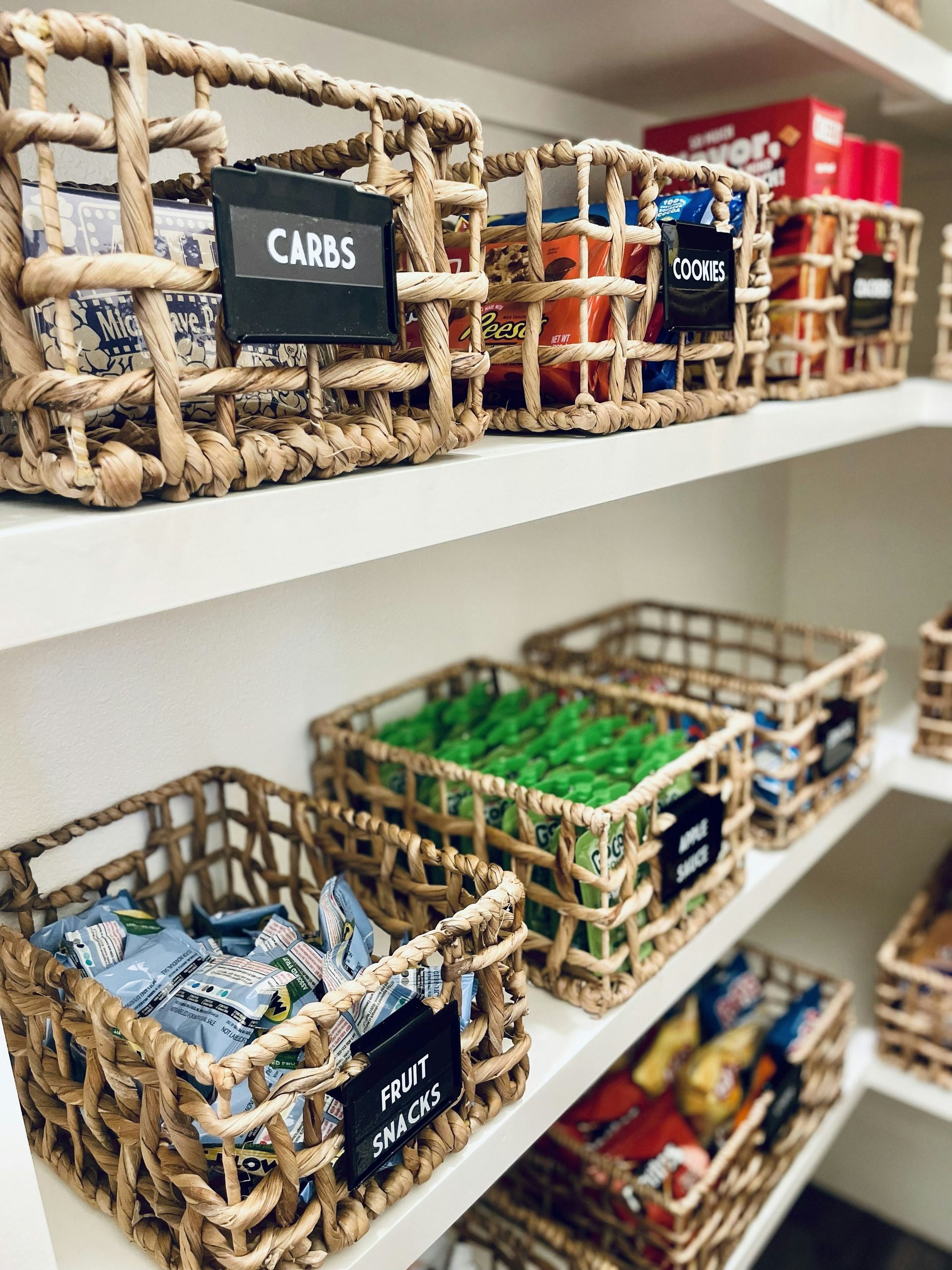 Woven baskets with labels on white pantry shelves filled with various snacks